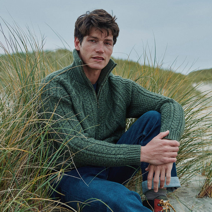 Man wearing a green knitted sweater sitting on a beach with grass.