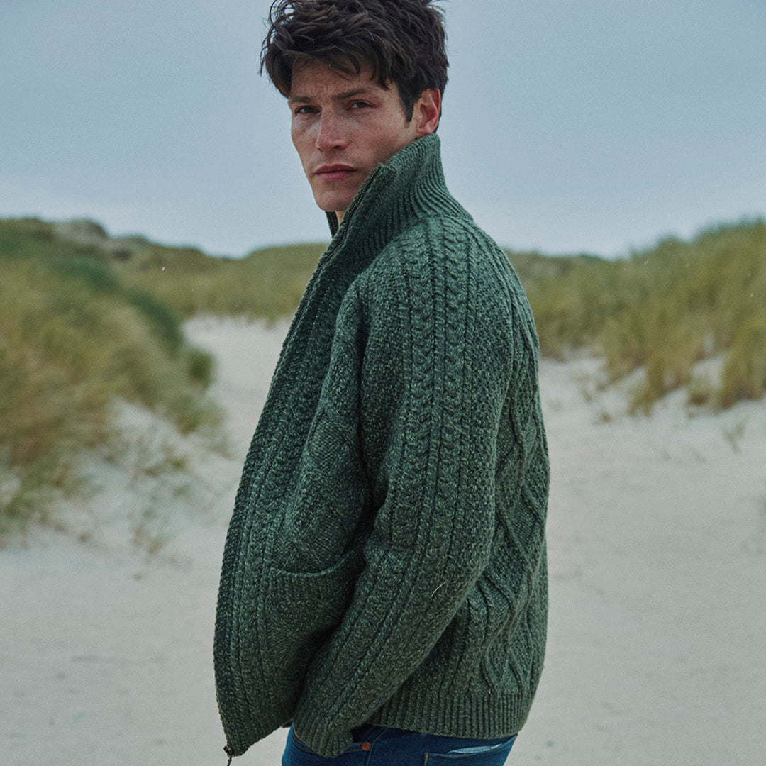 Man wearing a green knitted sweater and blue jeans on a beach, with 'aran woollen mills' branding.