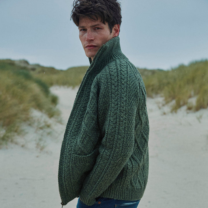 Man wearing a green knitted sweater and blue jeans on a beach, with 'aran woollen mills' branding.