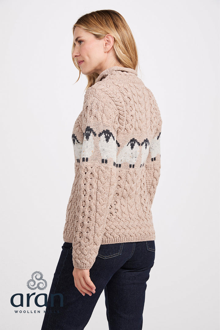 A woman wearing a beige Aran cable-knit sweater with a black and white sheep pattern across the back, paired with dark blue jeans, shown from a side-back angle against a light gray studio background.